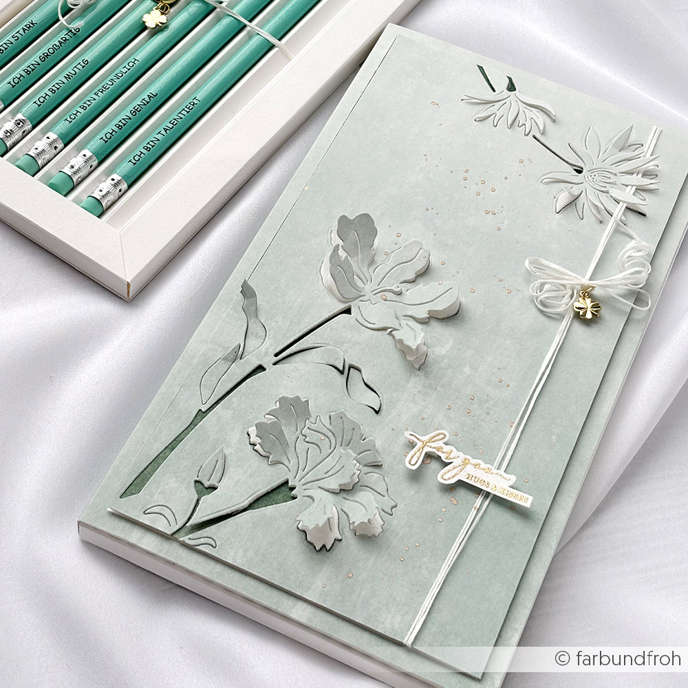 Ein pastellgrünes Blumenjournal mit geprägten Blumen, hergestellt mit Alexandra Renke Designpapieren und einer Motivstanze Negativ-Blume 5, liegt neben einer Schachtel mit grünen Bleistiften auf weißem Stoff.