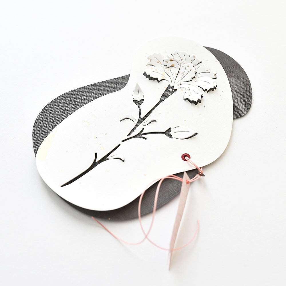 Scherenschnittblume und Knospen, hergestellt mit Alexandra Renke Motivstanze Negativ-Blume 6 auf geschichtetem weißem und grauem Designpapier, rosa Schnur befestigt, ausgestellt vor einem sauberen weißen Hintergrund.