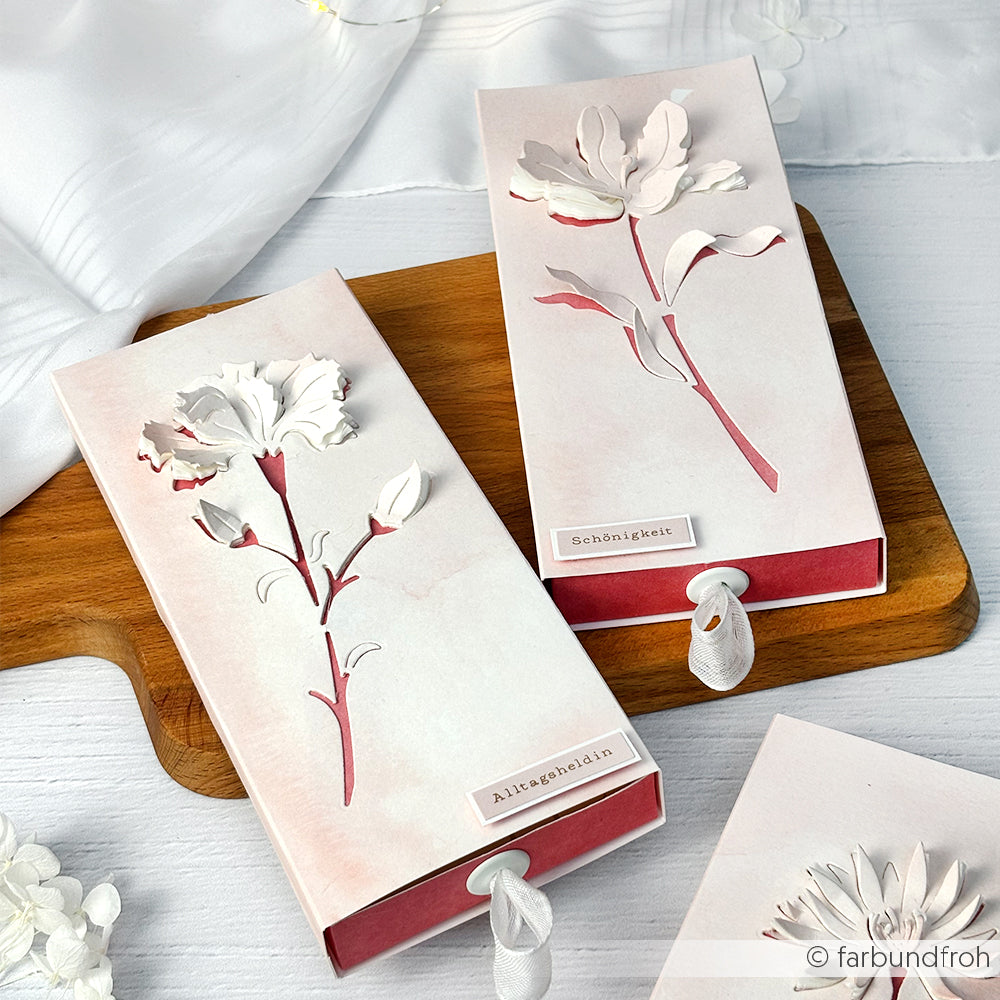 Zwei elegante Geschenkboxen von Alexandra Renke mit weißem und rosafarbenem Blumendesign auf hochwertigen Designpapieren, die mit DieCut Systemen gefertigt wurden, stehen auf einem Holzbrett mit sichtbaren Öffnungsbändern.