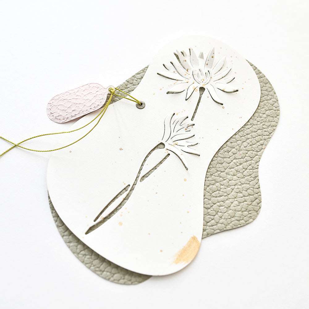 Weiße Karte von Alexandra Renke mit DieCut Systeme Blumenausschnitten auf strukturiertem grauem Leder, gebunden mit grünem Band und einem kleinen rosa Lederanhänger.