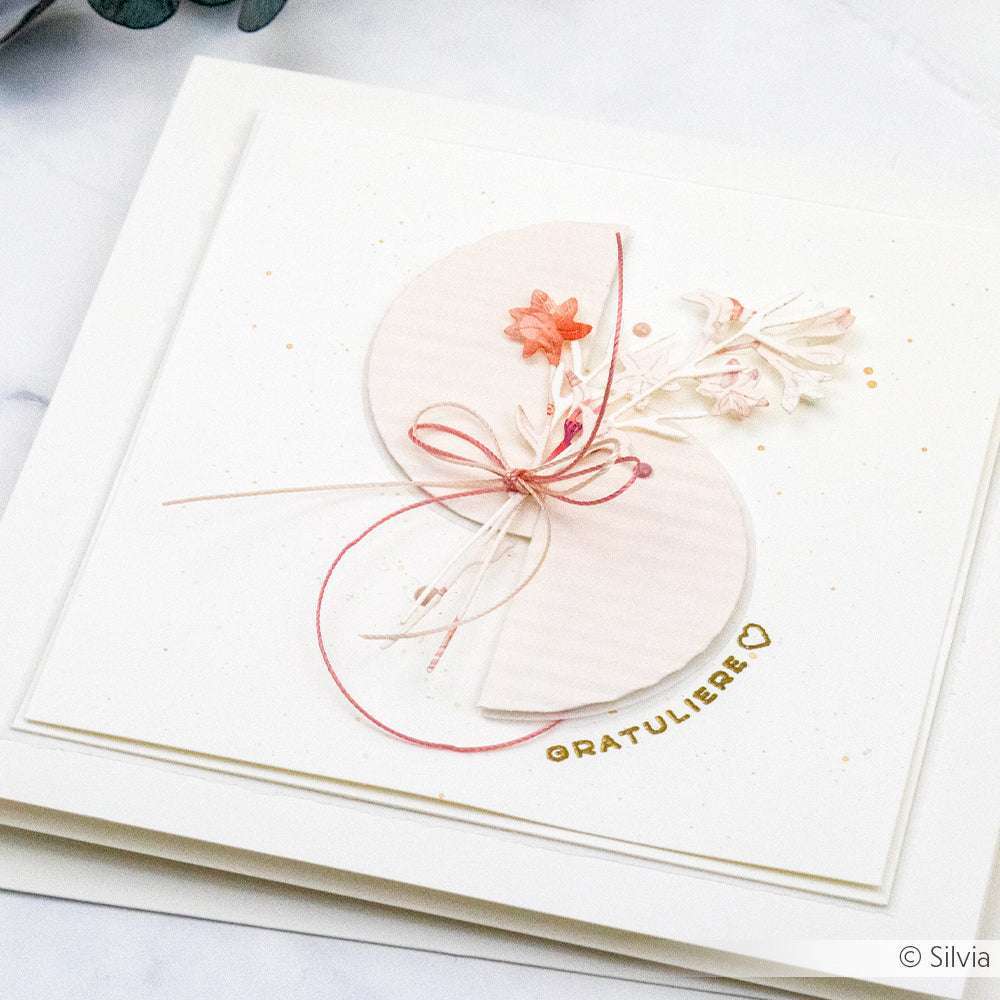 Elegante handgefertigte Karte von Alexandra Renke mit rosafarbenen Blüten aus Designpapieren, einer Schleife aus Garn und dem goldenen Text FÉLICITATIONS auf weißem Hintergrund. Mit zarten Blüten, die mit dem Kleine Kelchblüten DieCut System gestanzt wurden.