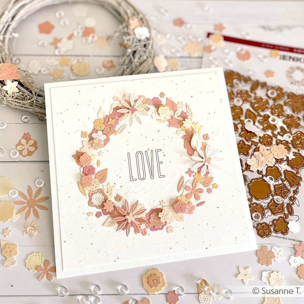 Quadratische Karte mit einem Blumenkranz und dem Wort LOVE, verziert mit Papierblumen aus Alexandra Renke Designpapier Stanzen und Motivstanze Blumenwiese, umgeben von passendem Bastelmaterial.