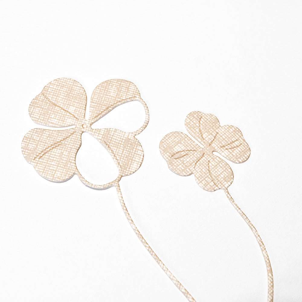 Zwei strukturierte, beigefarbene vierblättrige Kleeblätter mit dünnen Stielen auf weißem Untergrund, hergestellt mit der Alexandra Renke Motivstanze Glücksklee Duo - ideal, um Ihren Designpapieren und kreativen Stanzsystemen Charme zu verleihen.