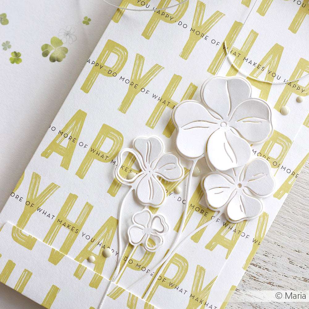 Weiße geprägte Blumen auf einer Karte, mit gelbem HAPPY-Text und zarten grünen Blumen im Hintergrund - gestaltet mit Alexandra Renke Designpapieren und dem Glücksklee Trio DieCut System für einen bezaubernden Lagenlook.