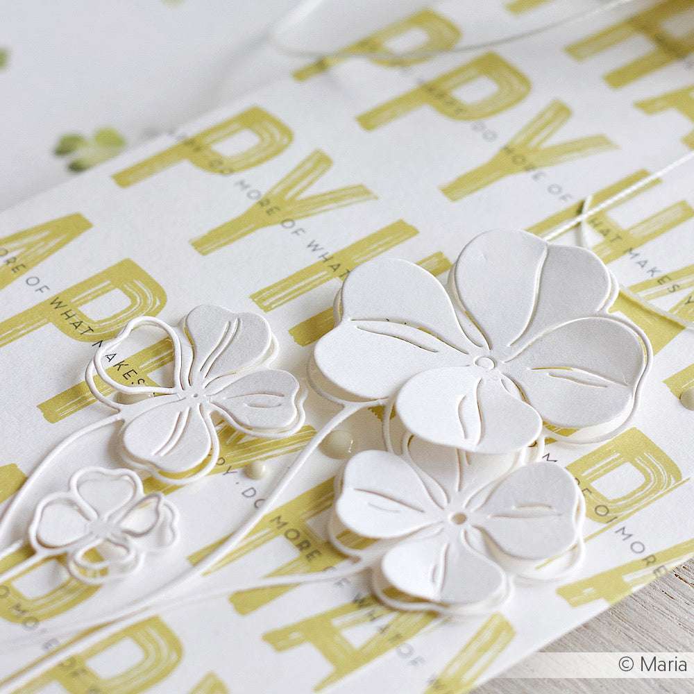 Weiße Scherenschnittblumen, die mit den Glücksklee Duo 2 Stanzformen von Alexandra Renke hergestellt wurden, sind auf einem gelb-weiß gemusterten Hintergrund arrangiert, wobei ein Teil des Textes zu sehen ist.