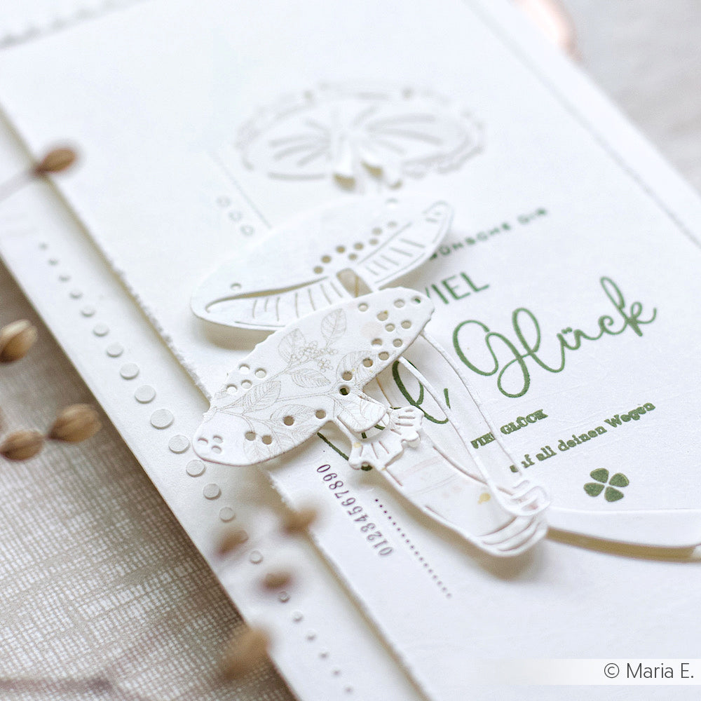 Nahaufnahme einer weißen, geprägten, handgefertigten Karte mit grünem Text und zarten ausgeschnittenen Regenschirmdekorationen, hergestellt mit Alexandra Renke Motivstanze Glückspilz Duo und hochwertigen Designpapieren.