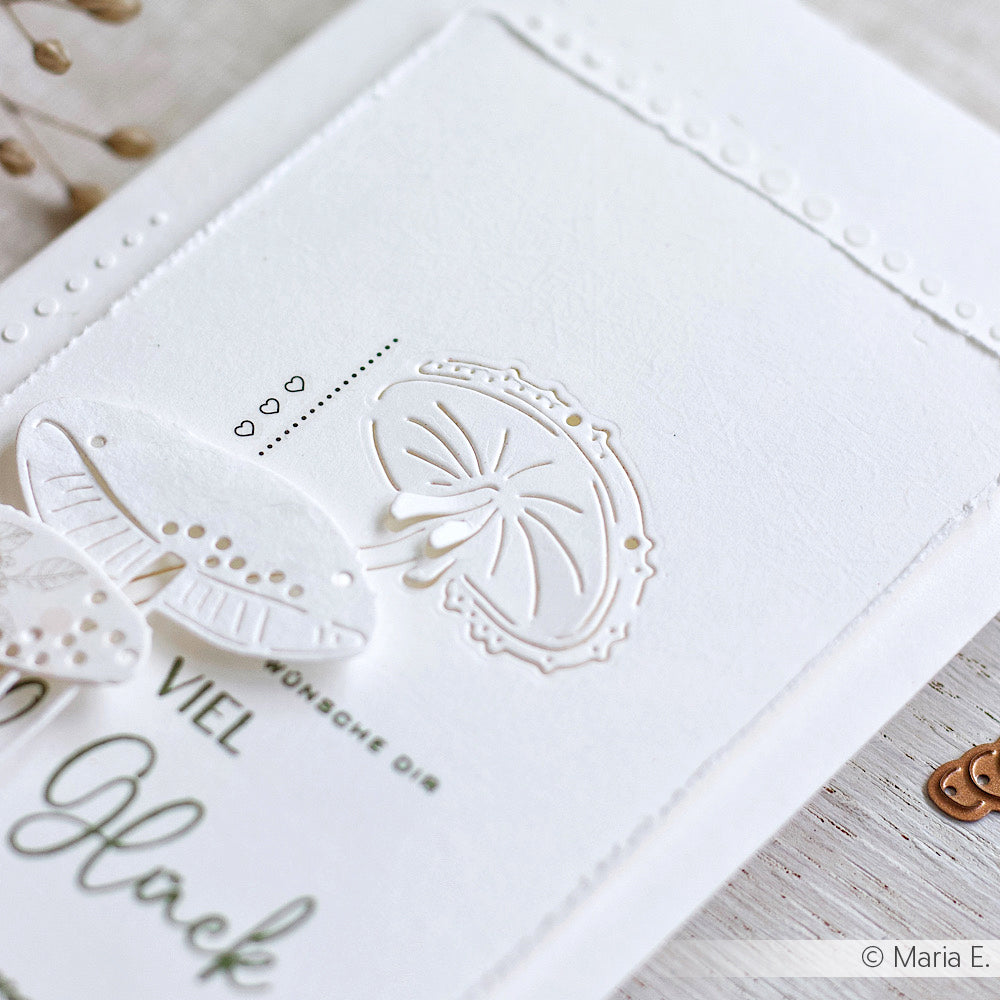 Weiße Prägekarte von Alexandra Renke mit zarten Schmetterlings- und Regenschirmmotiven, kleinen Herzen und deutschem Text auf hellem Hintergrund, hergestellt mit einer Stanzschablone für präzise Details.