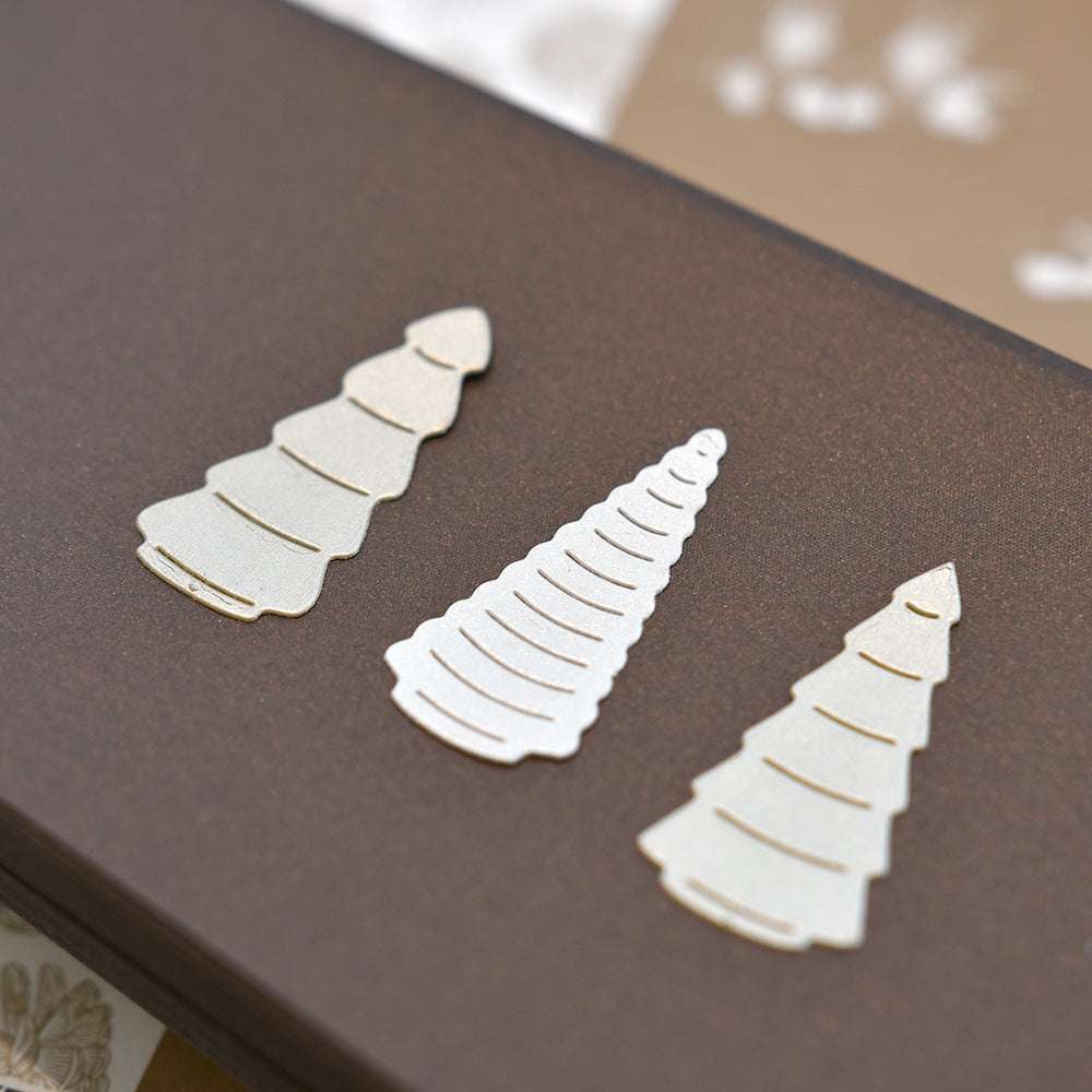 Drei metallische baumförmige Büroklammern von Alexandra Renke, inspiriert von der Motivstanze Abstraktes Baum Trio, ruhen auf einer braunen Oberfläche.