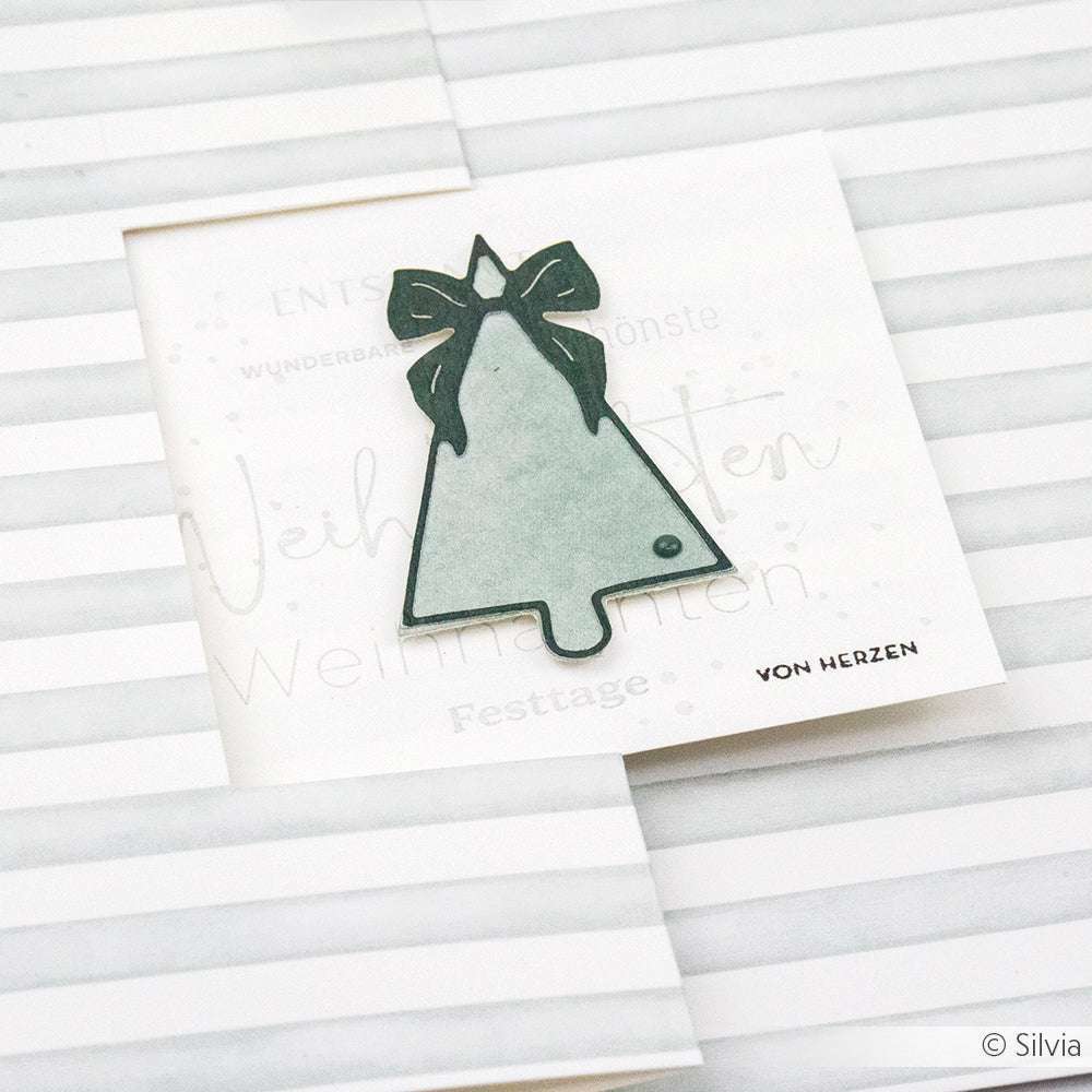 Die minimalistische Weihnachtskarte von Alexandra Renke zeigt einen schlichten Baum aus der Motivstanze Tanne und einer Schleife vor einem gestreiften Hintergrund aus ihren eleganten Designpapieren.