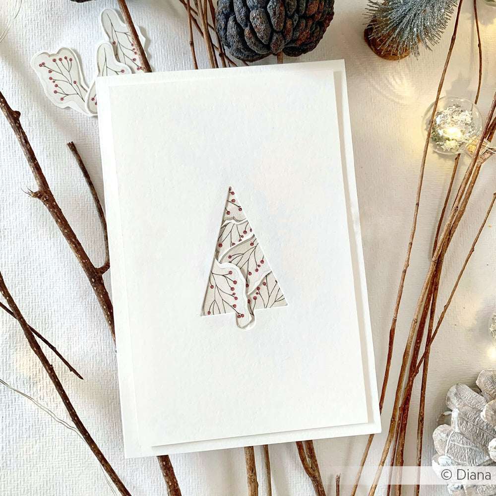 Weiße Karte mit einem baumförmigen Ausschnitt aus der Alexandra Renke Motivstanze Tannen Etikett, der ein florales Muster zeigt und mit Zweigen, Tannenzapfen und Lichterketten akzentuiert ist - ideal für Projekte mit Stanzsystemen oder Dies für Designpapiere.