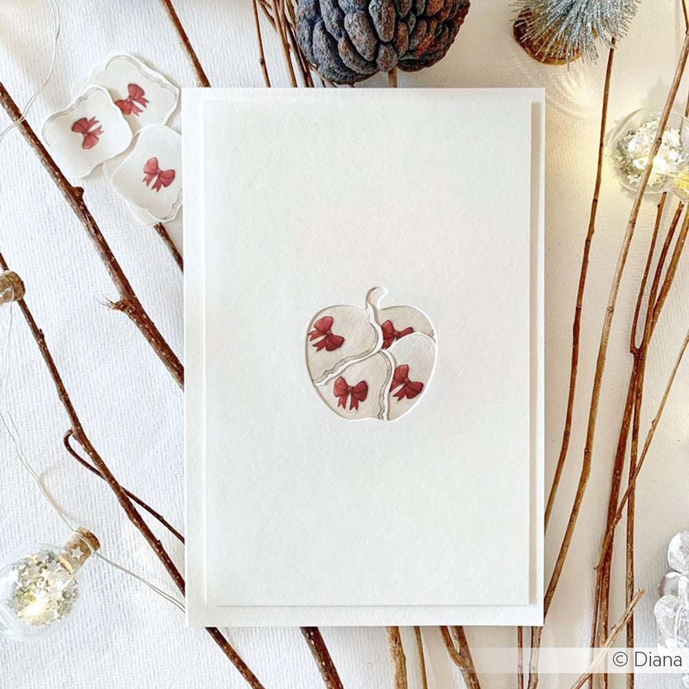 Weiße Karte mit einem Apfelausschnitt aus der Motivstanze Apfel Etikett Duo von Alexandra Renke, verziert mit roten Blumen, Zweigen, einem Tannenzapfen und Lichterketten. Ideal für kreative Projekte mit Designpapiere 200g oder Dies für Stanzsysteme.