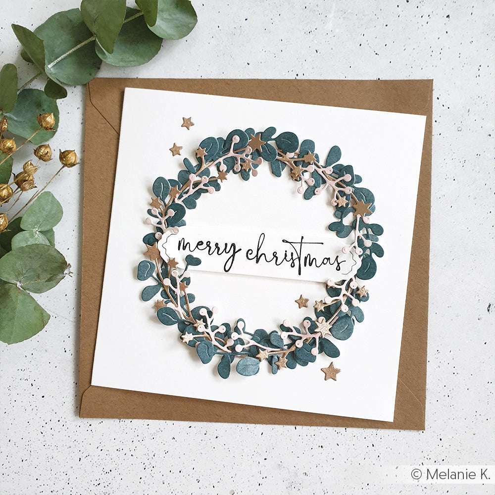 Quadratische Weihnachtskarte von Alexandra Renke mit grünem Kranz und dem Text "Frohe Weihnachten", hergestellt aus 200g Designpapier. Präsentiert auf einem braunen Umschlag, akzentuiert mit Grünzeug in der Nähe.