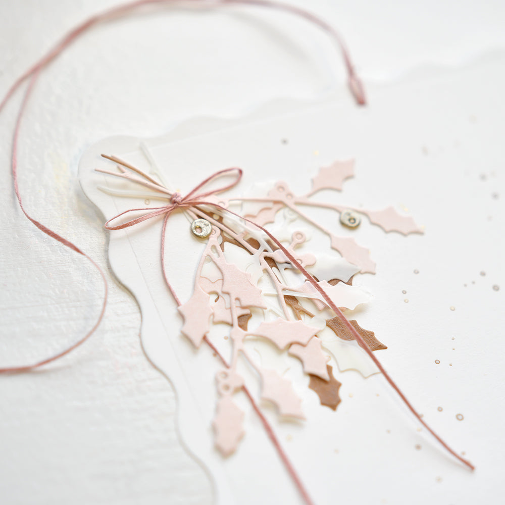 Zarte rosa und weiße Stechpalmenblätter, gefertigt mit der Alexandra Renke Motivstanze Ilex Trio, gebunden mit einer rosa Schnur auf einer weißen Karte mit Wellenschliff.