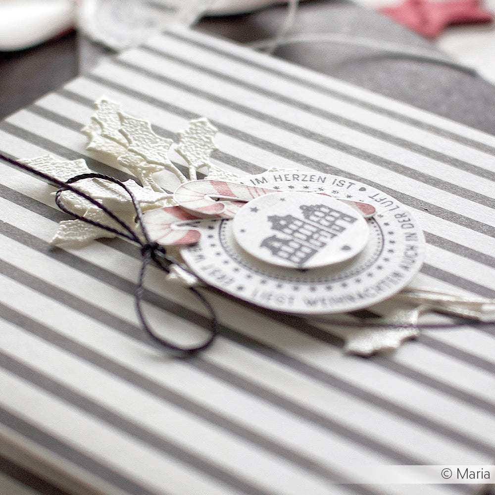 Nahaufnahme eines gestreiften Geschenkkartons, der mit festlichen Papierausschnitten, die mit Alexandra Renke DieCut Systemen hergestellt wurden, und einem runden Weihnachtsornament verziert ist.
