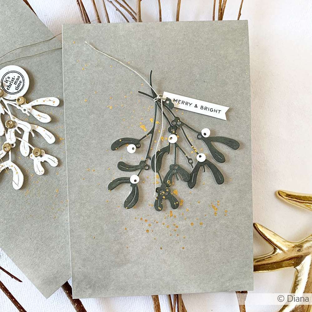 Handgemachte Karte mit Alexandra Renke Motivstanze Mistelzweig Trio, Goldspritzern und einem Merry & Bright Tag auf grauem Designpapier.