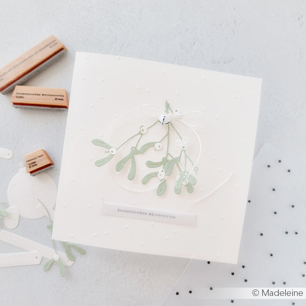 Eine handgefertigte weiße Karte mit zartgrünem Mistelzweig und einer kleinen Schleife, hergestellt mit Alexandra Renke's Motivstanze Mistelzweig Trio, wird inmitten von Alexandra Renke Stanzsystemen und Designpapieren präsentiert.