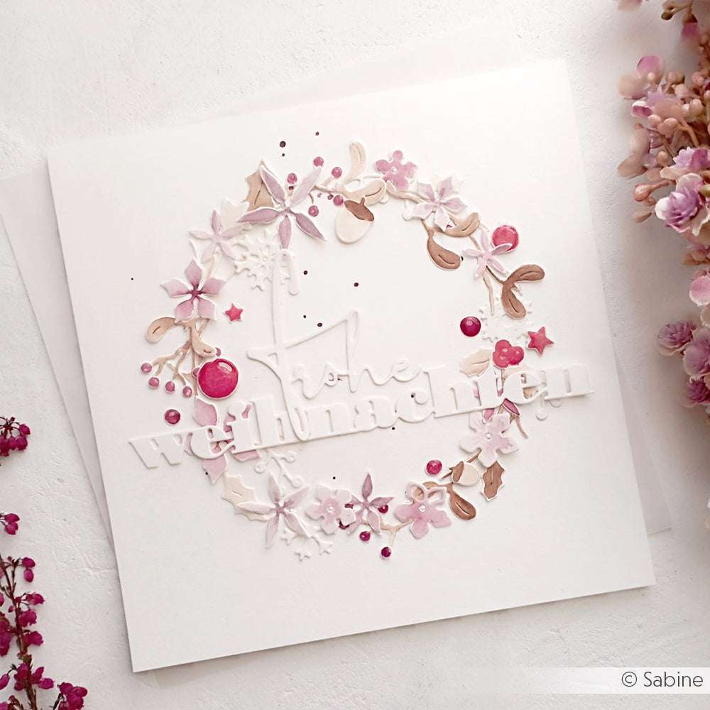 Handgefertigte Karte mit Alexandra Renke Designpapieren, die einen zarten rosa Blumenkranz aus der Motivstanze Weihnachtliche Blumensammlung und den erhabenen weißen Schriftzug Frohe Weihnachten" zeigt.
