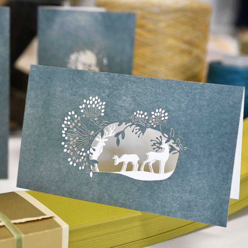 Eine blaue Grußkarte von Alexandra Renke mit einem lasergeschnittenen Hirsch- und Baummotiv, das mit DieCut Systemen hergestellt wurde, auf gelben Umschlägen.