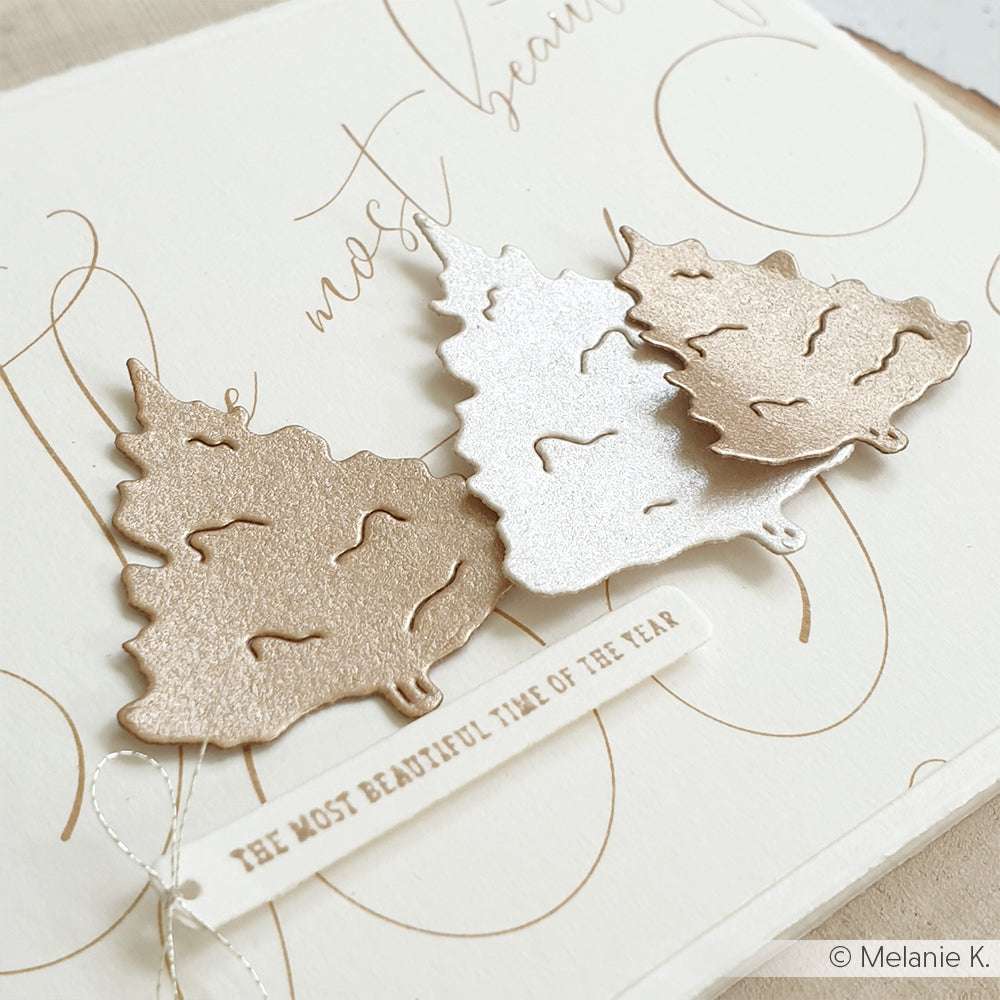 Drei Metallic-Papier-Weihnachtsbäume, hergestellt mit dem Alexandra Renke Motivstanze Tannen Trio, glänzen auf einer cremefarbenen Karte mit Schriftzug und einem Anhänger mit der Aufschrift "Die schönste Zeit des Jahres".