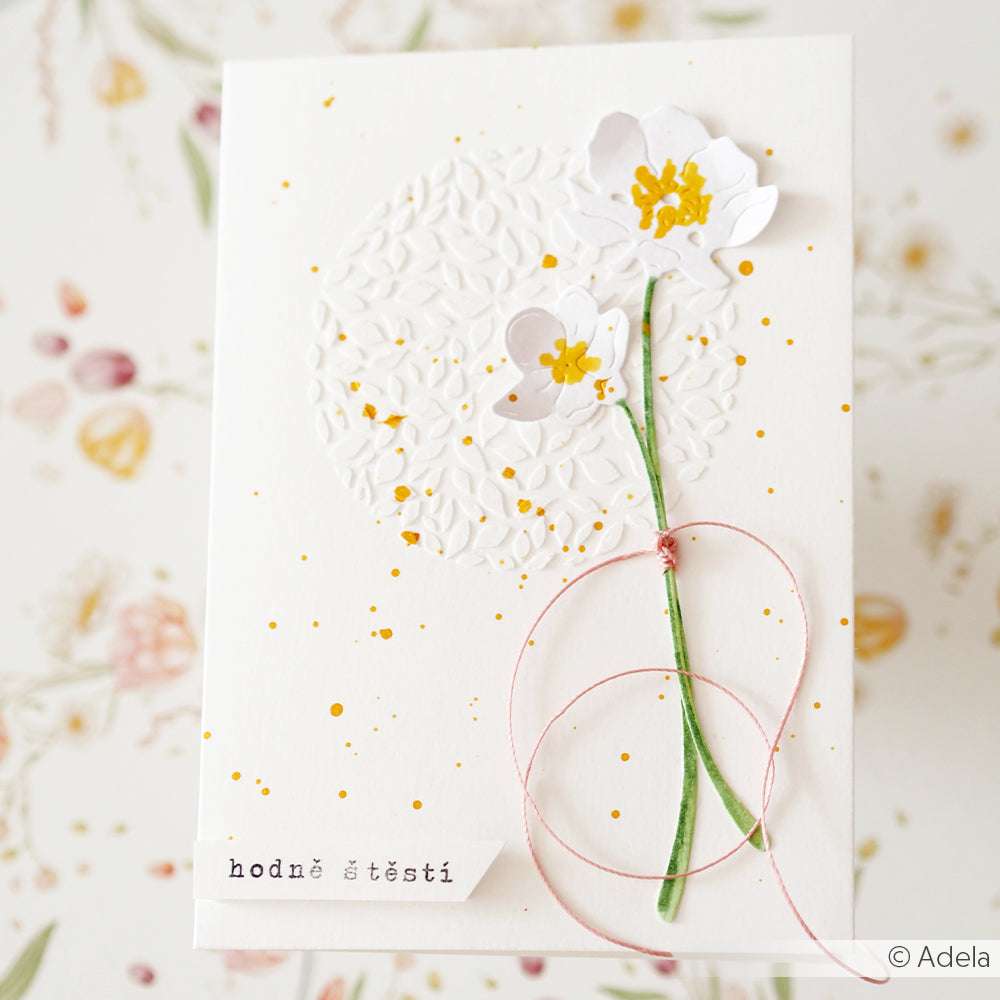 Eine weiße Karte von Alexandra Renke mit geprägter Textur, zwei weißen Blumen, rosa Schnur, dem tschechischen Text "hodně štěstí" und eleganten Details, die mit Deep Edge Motivstanzen hergestellt wurden.