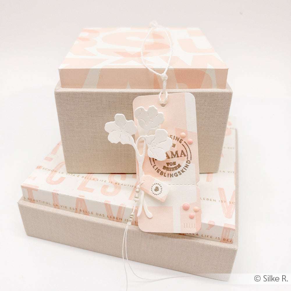 Mit Alexandra Renke Motivstanze Feines Blüten Duo gestalteter Geschenkanhänger mit weißem Blumendekor und "Mama"-Text auf gestapelten pastellrosa und beigen Geschenkkartons.