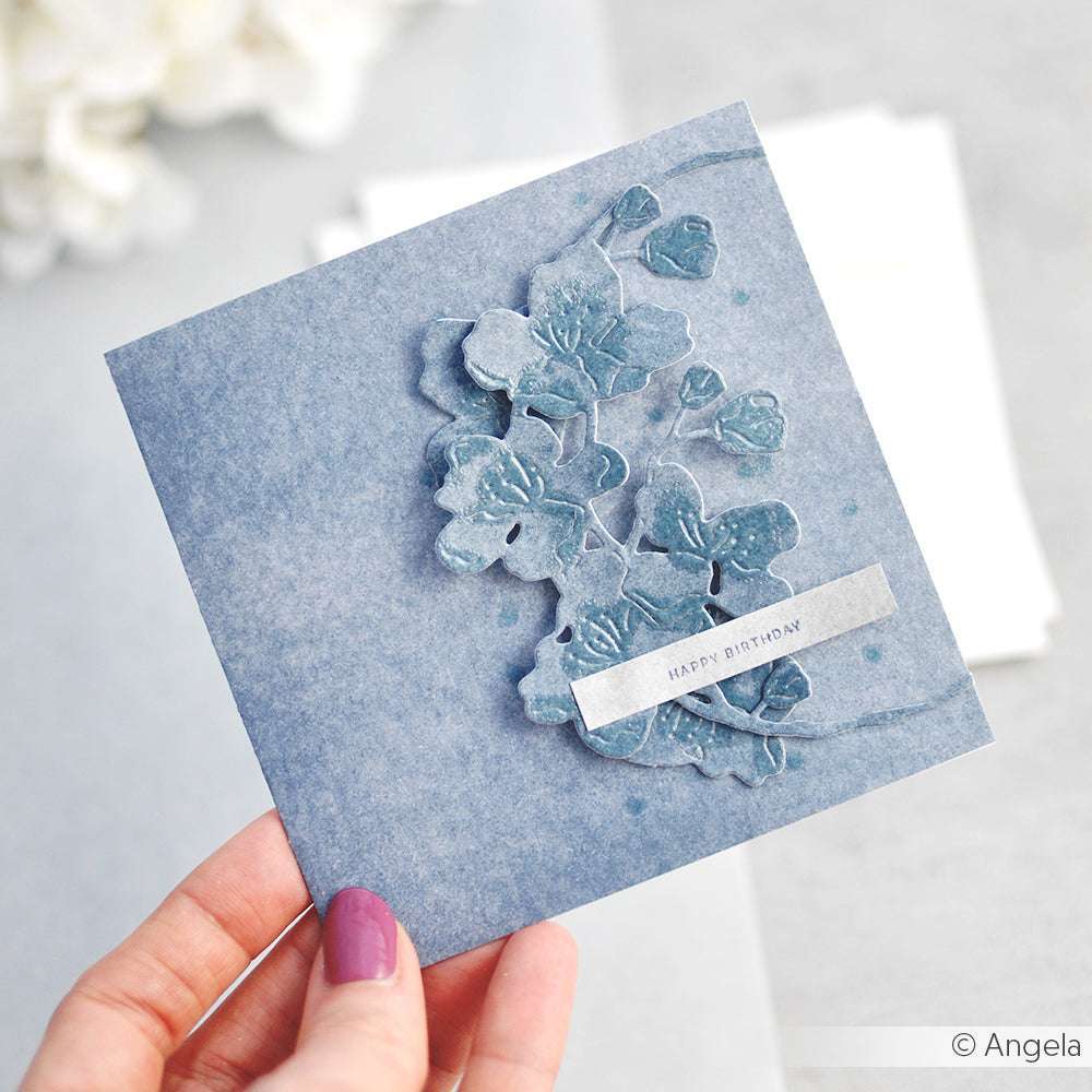 Eine Hand hält eine blaue Blumenkarte "Happy Birthday", verziert mit geprägten Blumen, die mit der Alexandra Renke Motivstanze Kirschblütenkranz hergestellt wurden.