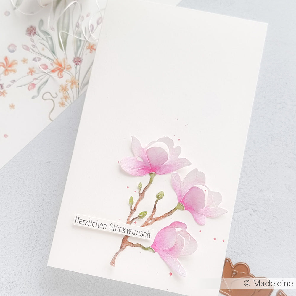 Weiße Karte mit rosafarbenen Aquarellblumen, hergestellt mit der Stanze Magnolienblüte von Alexandra Renke, mit dem deutschen Text "Herzlichen Glückwunsch" auf hellem Hintergrund.