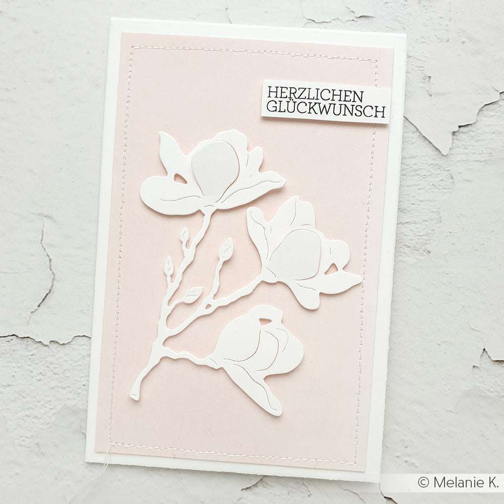 Handgefertigte Karte von Alexandra Renke mit weißen Blumenausschnitten, die mit Deep Edge Motivstanzen auf einem rosafarbenen Hintergrund hergestellt wurden, mit dem Vermerk "Herzlichen Glückwunsch" oben rechts.