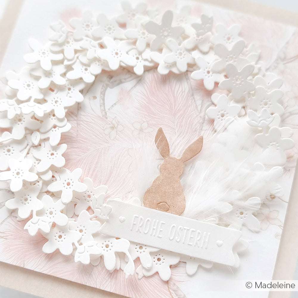 Weißer Blumenkranz mit einem kleinen braunen Hasen und dem Spruchband "Frohe Ostern!" auf pastellrosa, mit zarten Akzenten, inspiriert von der Motivstanze Vergissmeinnicht von Alexandra Renke, für einen bezaubernden Frühlingslook.