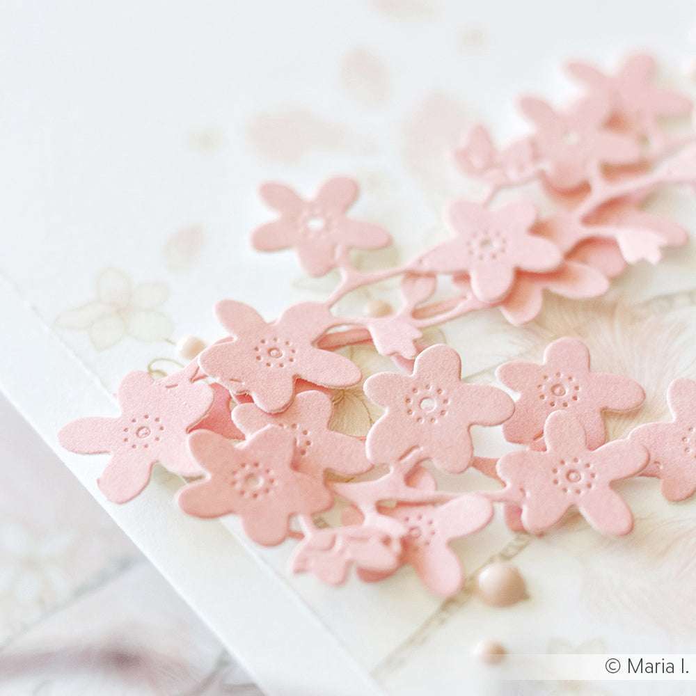 Nahaufnahme von zarten, hellrosa Papierblumen, die mit der Alexandra Renke Motivstanze Vergissmeinnicht hergestellt wurden, arrangiert auf einem weichen, floral gemusterten Hintergrund.