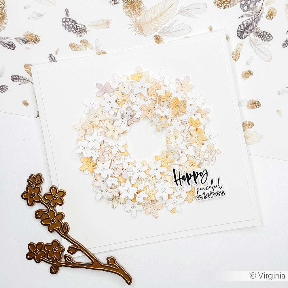 Eine handgefertigte Karte mit einem Kranz aus kleinen pastellfarbenen Blumen und dem Text "Happy peaceful wishes", neben einer Alexandra Renke Deep Edge Motivstanze Vergissmeinnicht.