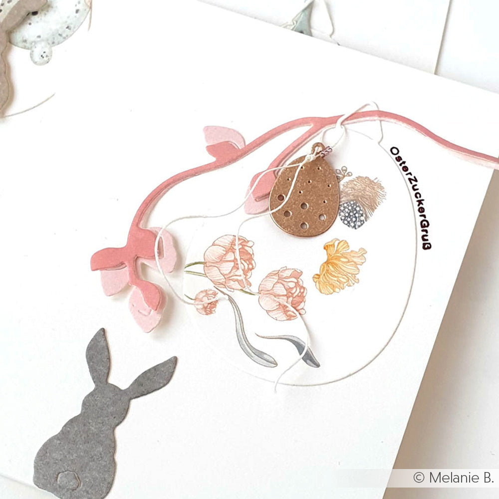 Eine handgefertigte Karte von Alexandra Renke mit einem grauen Hasen, einem rosafarbenen Zweig, Blumendrucken und einem kleinen braunen Anhänger auf weißem Hintergrund - gefertigt mit Deep Edge Motivstanzen für filigrane Details.