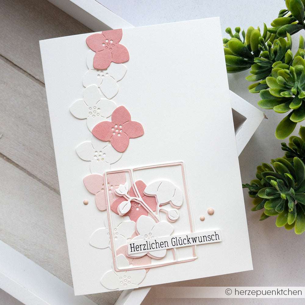 Eine weiße Karte von Alexandra Renke mit rosa und weißen Blumenverzierungen, die mit Deep Edge Motivstanzen hergestellt wurden, sowie dem deutschen Text "Herzlichen Glückwunsch" auf der Vorderseite.