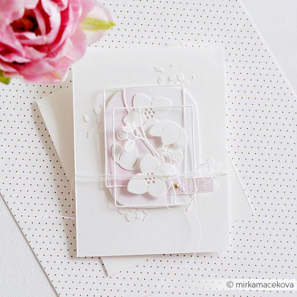Eine handgefertigte, geschichtete, weiße Blumen-Grußkarte, die mit der Alexandra Renke Blumenrahmen-Stanze hergestellt wurde, neben einer rosa Rose auf gepunktetem Hintergrund.
