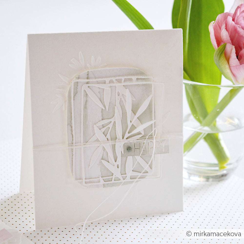 Eine handgefertigte weiße Grußkarte mit einem botanischen Design von Alexandra Renke, hergestellt mit der Motivstanze Gräserrahmen, steht neben einer Vase mit grünen Blättern und einer rosa Blüte.