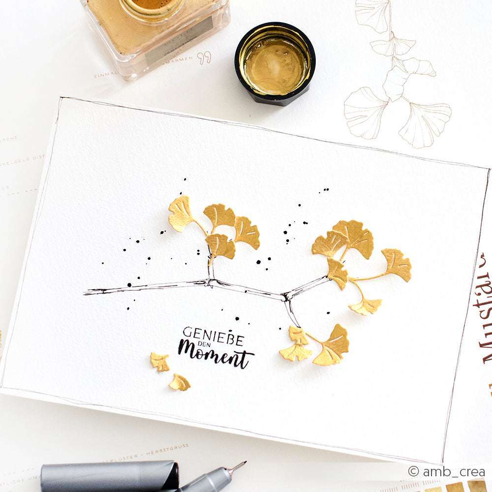 Eine Skizzenbuchseite mit goldenen Ginkgoblättern aus der Alexandra Renke Motivstanze Mini Ginkgozweige an einem Zweig und darunter handschriftlich "Genieße den Moment", gefertigt auf elegantem Alexandra Renke Designpapier 200g.