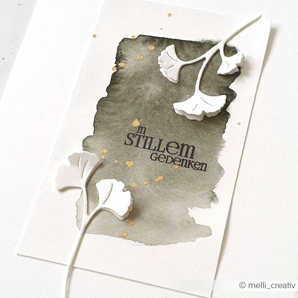 Karte mit schwarzem Aquarell-Hintergrund, Goldspritzern, weißen Blattausschnitten aus der Alexandra Renke Mini Ginkgozweige Stanze und dem deutschen Text "In stillem Gedenken".