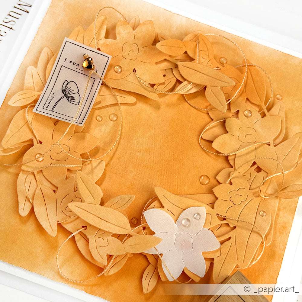 Ein handgefertigter Papierblumenkranz, hergestellt mit Alexandra Renkes Motivstanze Cymbidium-Kranz und PAPPcut®ies, wird auf einem warmen orangefarbenen Hintergrund mit einer beigefügten Karte und einer goldenen Anstecknadel präsentiert.