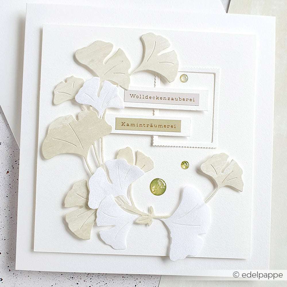 Handgemachte Karte mit geschichteten Ginkgoblättern aus der Alexandra Renke Motivstanze Ginkgo-Kranz, zwei deutschen Etiketten und drei grünen Edelsteinen auf weißem Hintergrund.