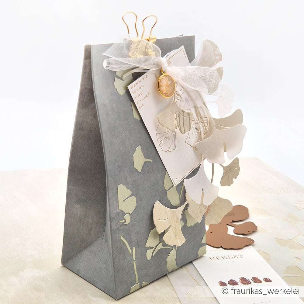 Eine graue Geschenktüte mit Ginkgoblättern aus Papier, die mit der Motivstanze Ginkgo-Kranz von Alexandra Renke, Geschenkband, Anhänger und einer goldenen Büroklammer verziert ist - perfekt für kreative Projekte mit DieCut Systemen.