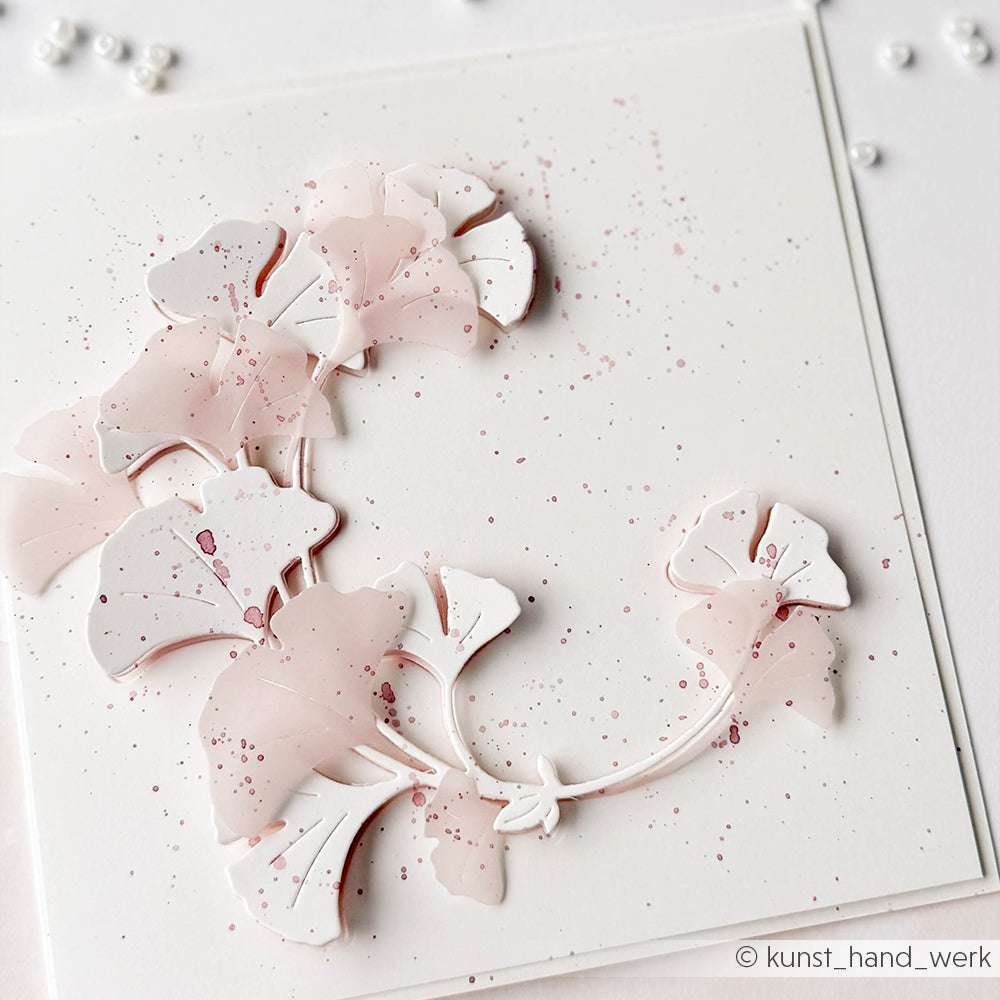 Zarte rosa und weiße Papierblätter, die mit der Alexandra Renke Ginkgo-Kranz-Stanze hergestellt wurden, sind in einem sanften Bogen auf einem gesprenkelten weißen Hintergrund angeordnet.
