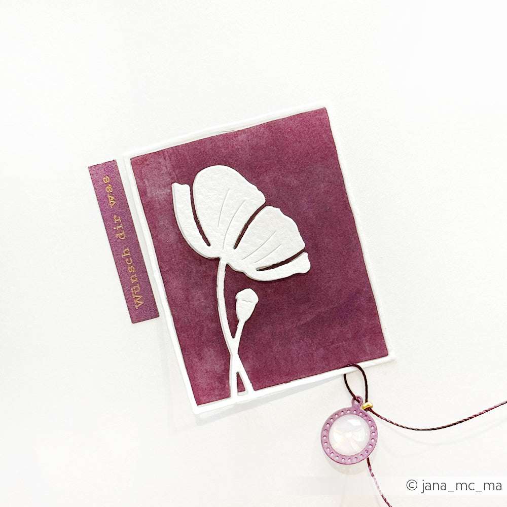 Handgefertigte Karte von Alexandra Renke mit weißer Blumensilhouette und dem Motivstanze Mohn im Rahmen auf kastanienbraunem Karton, verziert mit einem runden Anhänger, der an einer Schnur befestigt ist.