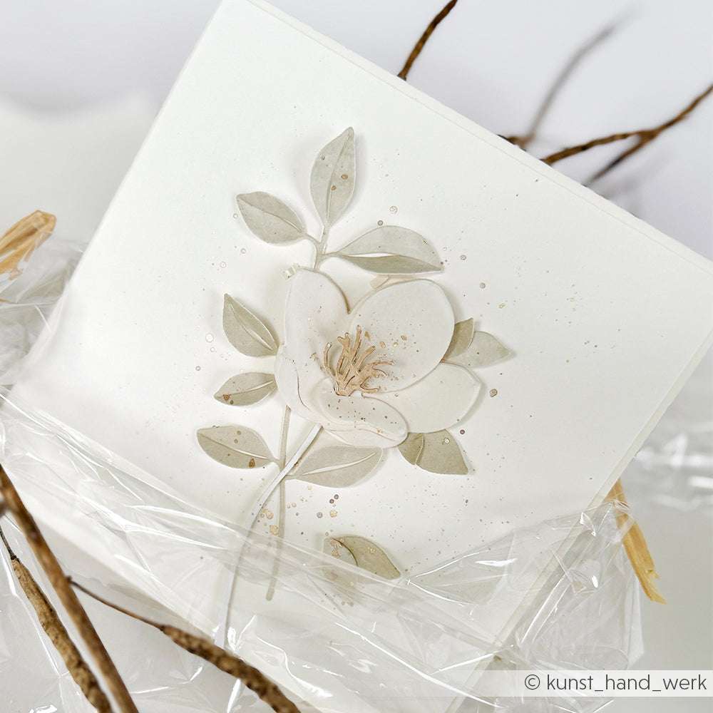 Handgefertigte Karte von Alexandra Renke mit weißer Blüte und Blättern mit zarten Deep-Edge-Stanzdetails, verpackt in klarem Plastik.