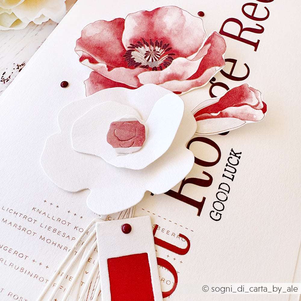 Nahaufnahme von roten und weißen Papierblumen auf einer strukturierten Karte mit dem Text "Good Luck", die mit Alexandra Renke-Produkten für dekorative Details gestaltet wurde.