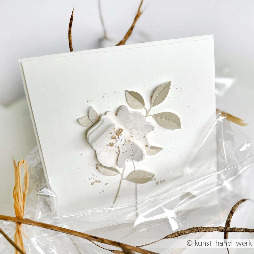 Die weiße Grußkarte von Alexandra Renke zeigt ein geprägtes florales Motiv aus der Motivstanze Zarte Formblüte, das auf klarem Kunststoff aufgebracht und mit strohhalmartigen Verzierungen akzentuiert ist.