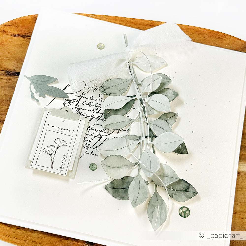 Handgemachte Karte mit Papierblättern aus der Alexandra Renke Motivstanze, einem kleinen Anhänger, Schrift und einer Tüllschleife auf weißem Hintergrund.