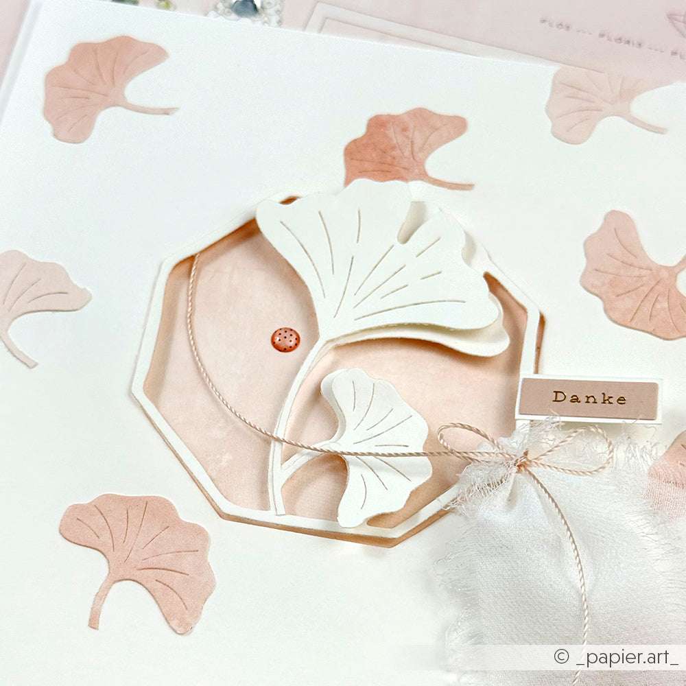 Handgefertigte Karte mit Ginkgo-Blattausschnitten aus der Alexandra Renke Motivstanze Ginkgo, mit einem kleinen Danke-Anhänger und zartrosa und weißen dekorativen Details.