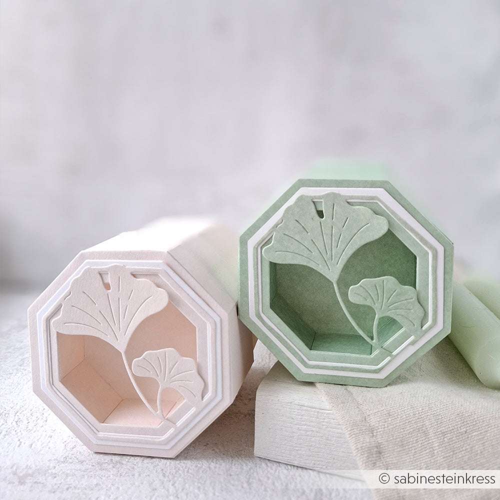 Zwei sechseckige pastellfarbene Schachteln mit Ginkgoblattmotiven, hergestellt mit der Alexandra Renke PAPPcut Motivstanze Ginkgo in Rosa und Grün, ruhen auf einer weißen Fläche.