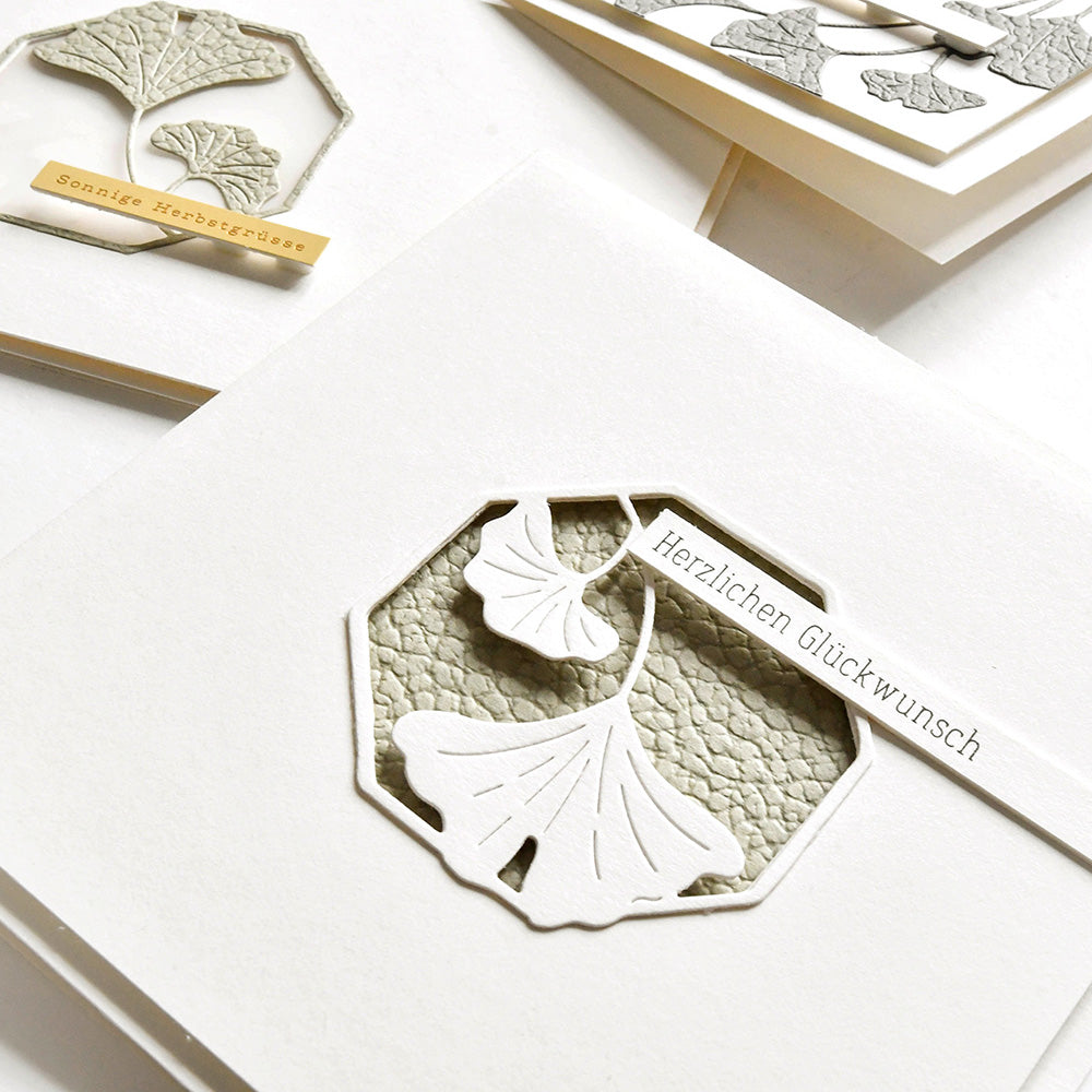 Handgefertigte weiße Karten mit Ginkgo-Blattausschnitten unter Verwendung der Ginkgo-Motivstanze von Alexandra Renke und deutschem Text, auf strukturiertem grauem Hintergrund.