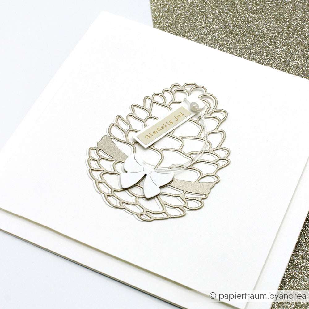 Handgemachte Karte von Alexandra Renke mit einer silbernen Tannenzapfen-Stanzung, einer weißen Schleife und einem "Glædelig jul"-Anhänger auf hochwertigem Designpapier, alles vor einem weißen Hintergrund.